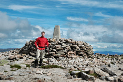 Picture of a walker in front of the summit cairn and trig point on Ben Macdui