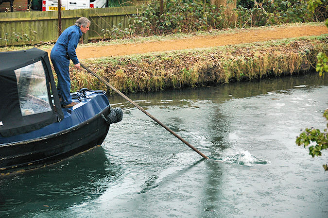 Picture of a man standing on the bow of a canal boat breaking ice on the water with a pole